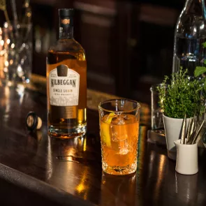 A glass of The Wheel with ice and lemon slice alongside bottle of killbeggan Whiskey and a plant pot