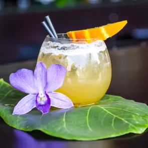 A glass of Mango Rum Margarita garnished with orange slice and two strawss with a purple flower on a leaf