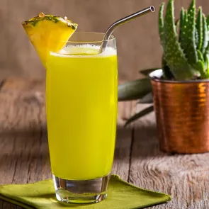 Glass of Midori® Cactus Juice with a  straw, garnished with a pineapple wedge on a green napkin, alongside a cactus plant.