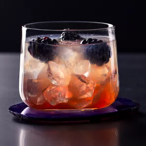 "A Glass of Zarza garnished with ice cubes and mulberry fruit on a  coaster